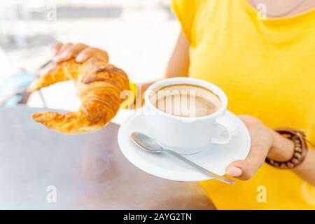 Glückliche Frau in Gelb, die im Café im Freien mit Croissants und Kaffee frühstücken kann Stockfoto