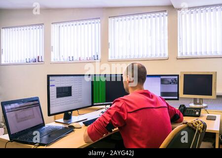 Hinter dem Schreibtisch eines Mitarbeiters sitzt ein Mann im Büro. Der Techniker überwacht das Computernetzwerk. Ein Spezialist arbeitet für mehrere Computer. Stockfoto