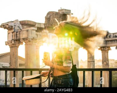 Schöne junge Frau, die bei Sonnenaufgang lange blonde Haare auf dem Forum Romanum schüttelt. Fröhlicher lächelnder Tourist mit Oldtimer-Kamera. Objektivflair. Rom, Italien Stockfoto
