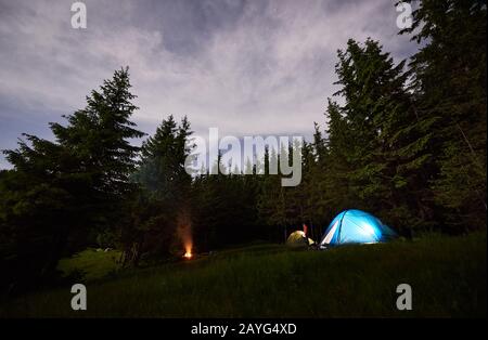 Abends im Wald campen. Zelte und ein brennendes Feuer unter grünen Tannen unter Sternenhimmel mit einem Dunst von Rauch. Person zwischen Zelten, in denen Lampen leuchten. Gesundes Lifestyle-Konzept Stockfoto