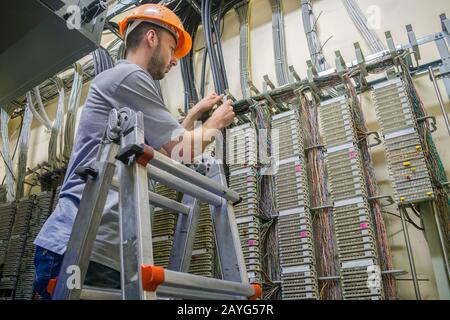 Ein Arbeiter, der auf der Treppe steht, pendelt Telefonleitungen. Der Techniker arbeitet im Serverraum des Rechenzentrums. Kabelverlegung im Telefonaustausch Stockfoto