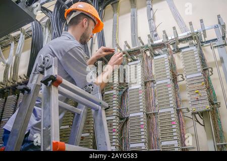 Der Techniker arbeitet im Serverraum des Rechenzentrums. Kabelverlegung im Telefonaustausch. Ein Arbeiter, der auf der Treppe steht, pendelt Telefonleitungen Stockfoto