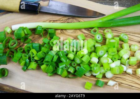 Nahaufnahme von isolierten, in Scheiben geschnittenen frischen Frühlingszwiebeln (Hengste) auf Holz-Oliven-Baum-Schneidebrett und japanischem Küchenmesser Stockfoto