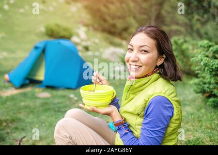 Morgens warmes Frühstück im Wald - Frau Wanderer, die beim Zelten in den Bergen warmes Essen in der Nähe des Zeltes isst Stockfoto