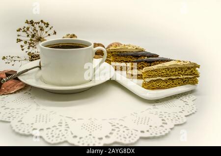 Eine weiße Tasse mit schwarzem Kaffee und einem silbernen Löffel, ein weißes Gericht mit Scheiben von Cremetakuchen auf einer weißen Papier-Öffnungs-Serviette Stockfoto