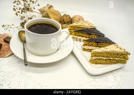 Weiße Tasse mit schwarzem Kaffee und einem silbernen Löffel, eine weiße Schale mit Scheiben von Cremetakuchen auf einer weißen PapieröffnungsServiette Stockfoto