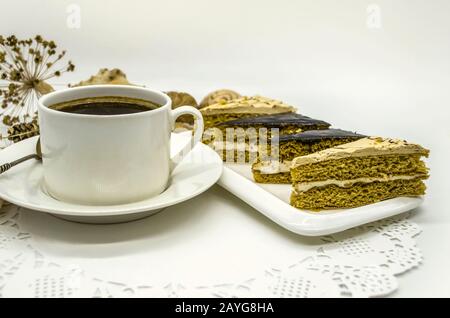 Trockene Blumen und ein Kiefernkegel hinter einer weißen Tasse schwarzen Kaffees und ein weißes Gericht mit Scheiben von Cremekuchen liegen auf einer weißen PapieröffnungsServiette Stockfoto