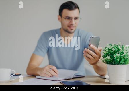 Modernes Smartphone in den Händen des Mannes. Ein vielbeschäftigter Büroangestellter erhält Nachrichten auf dem Handy, während er im Tagebuch Notizen macht, sitzt gegen gemütliche Interi Stockfoto