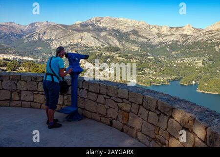 Dame, die durch das Teleskop schaut, die uralte Berg-Zitadelle von Guadalest, in der Nähe von Benidorm, an der Costa Blanca, Spanien mit dem Reservoir in der Ferne Stockfoto