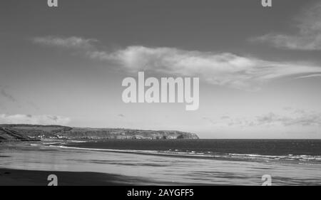 Whitby Beach. Wellen rollen auf einen nassen Strand. In der Ferne befindet sich eine Landzunge und ein Gebäudehaufen. Ein Himmel mit Wolken ist oben. Stockfoto