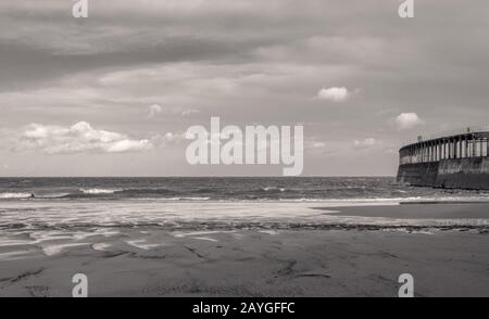 Whitby Beach. Wellen rollen sich auf den vom Wind geformten nassen Sand ein. Ein Pier erstreckt sich bis zum Meer und ein bewölkter Himmel ist über. Stockfoto