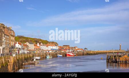 Ein Panorama auf den Außenhafen von Whitby. Die Gebäude führen an der Küste entlang, und Boote werden zu einem Kai hin vermauert. Eine Anlegestelle und ein Leuchtturm befinden sich in der Ferne. Stockfoto