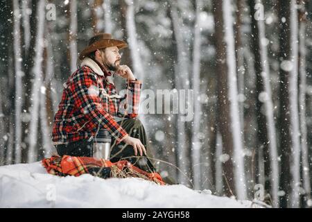 Nachdenklicher männlicher Wanderer in karierter Jacke, der auf einem kleinen verschneiten Waldhügel sitzt und in die Ferne blickt Stockfoto