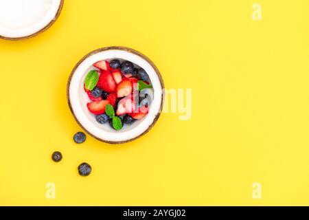 Direkt über dem Blick auf die Kokosschale mit frischen reifen Beeren auf hellgelbem Hintergrund mit Kopierraum Stockfoto