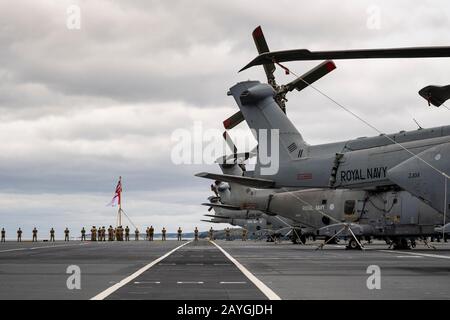 Die Schwänze der aufgereihten Merlin-Hubschrauber der Royal Navy an Bord des Trägers "HMS QUEEN ELIZABETH" mit Royal Marines, die die Schiene im Hintergrund säumen. Stockfoto