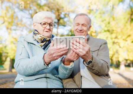 Senioren-Paar sitzt auf einer Bank im Herbstpark und verwendet digitales Tablet Stockfoto