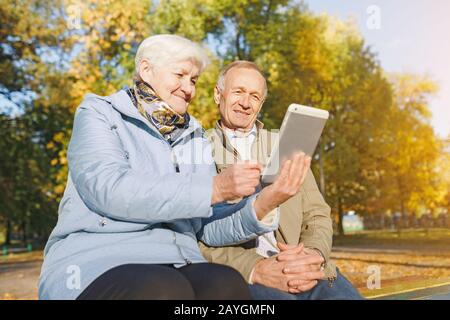 Senioren-Paar sitzt auf einer Bank im Herbstpark und verwendet digitales Tablet Stockfoto