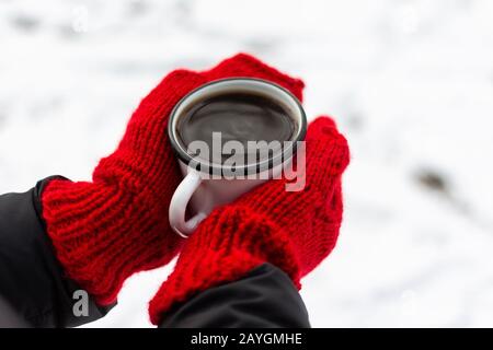 Weibliche Hände in rot gestrickten Fäusten, in den Händen eines eisernen Becher mit Kaffee oder Tee auf dem Hintergrund von weißem Schnee Stockfoto