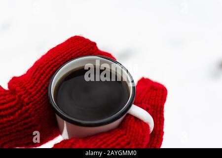 Weibliche Hände in rot gestrickten Fäusten, in den Händen eines eisernen Becher mit Kaffee oder Tee auf dem Hintergrund von weißem Schnee Stockfoto