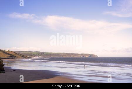 Whitby Beach. Wellen rollen auf einen nassen Strand, während ein Paar seinen Hund spazieren geht. Eine Landzunge liegt in der Ferne und ein Himmel mit Wolken darüber. Stockfoto