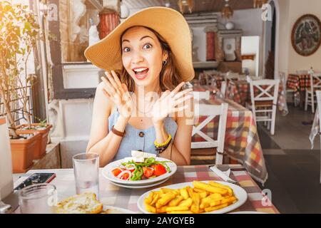Glückliche asianerin in großem Hut, die im Restaurant mit griechischem Salat Horiatiki gegessen hat. Konzept der griechischen Küche Stockfoto