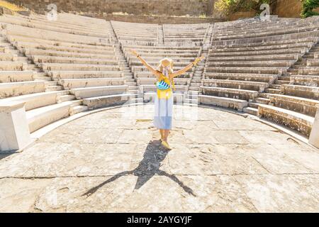 Touristen asiatischer Mädchen, die in den Ruinen einer antiken Akropolis oder dem Amphitheater mit einer griechischen Flagge unterwegs sind. Aktive junge Reisende und Studenten Konzept