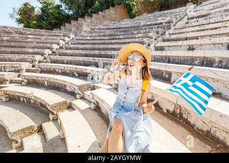 Touristen asiatischer Mädchen, die in den Ruinen einer antiken Akropolis oder dem Amphitheater mit einer griechischen Flagge unterwegs sind. Aktive junge Reisende und Studenten Konzept Stockfoto