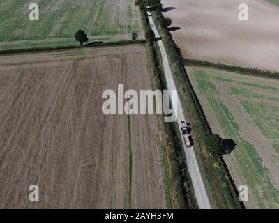 Luftansicht eines Traktors und Anhängers auf einer langen geraden Straße mit Hecken und gepflügten Feldern auf beiden Seiten Stockfoto