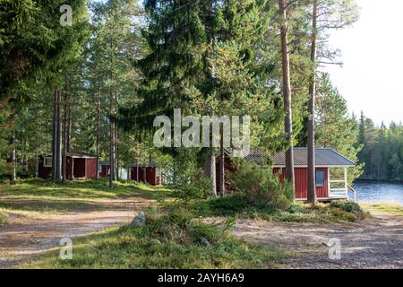 Rote finnische Holzhütten in traditionellen Hütten in grünem Kiefernwald in der Nähe des Flusses. Ländliche Architektur Nordeuropas. Holzhäuser im Camping auf der Sonne Stockfoto