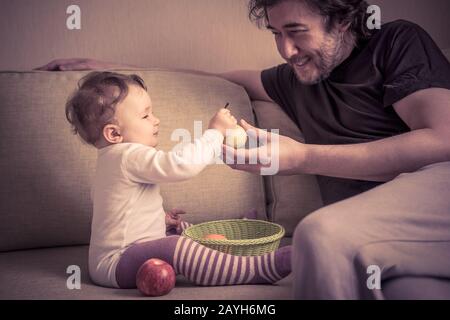 Glückliches Baby-Mädchen spielt mit Früchten bei ihrem Vater zu Hause. Das einjährige Kind hält eine Birne in der Hand. Stockfoto