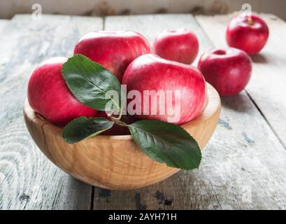 Mehrere rote Äpfel mit grünen Blättern liegen in einer Holzschale nahe0oben auf dem alten Holztisch Stockfoto