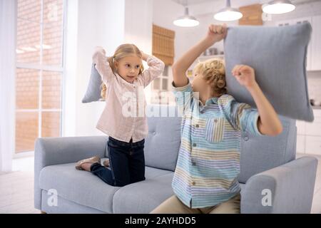 Zwei Kinder zahlen auf dem Sofa und schlagen sich mit Kopfkissen Stockfoto