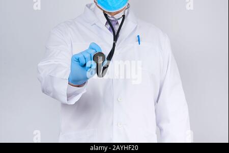 Doktor in einem weißen Mantel und Krawatte hält in der Hand ein schwarzes Phonendoskop, das blaue sterile Handschuhe trägt, weißer Hintergrund Stockfoto