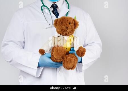 Kinderarzt in weißem Mantel, blaue Latexhandschuhe halten einen braunen Teddybären mit gelbem Band an einem Pullover, Konzept des Kampfes gegen Kinderkäuze Stockfoto