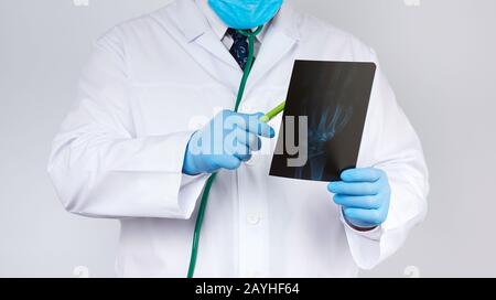 Doktor in einem weißen Mantel und blaue Latexhandschuhe halten eine Röntgenaufnahme der Hand eines Mannes und führen eine visuelle Untersuchung durch, weißer Hintergrund Stockfoto