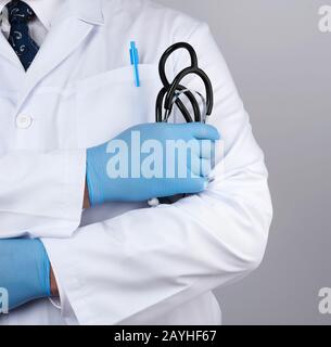 Ein ausgewachsener männlicher Arzt in einem weißen medizinischen Mantel steht und hält ein schwarzes Stethoskop auf weißem Grund, nah oben Stockfoto