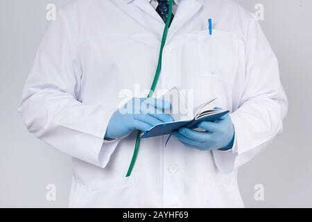 Doktor in einem weißen Mantel und blauen Handschuhen hält ein blaues Papier-Notizbuch für Notizen, Stethoskop hängt an seinem Hals, weiße medizinische Kulisse Stockfoto