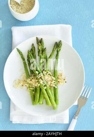 Lebhafter grüner, gedämpfter Spargel mit Parmesan, Petersilie und roten Pfefferflocken-Brotkrümeln bestreut. Auf weißer Platte auf blauem Hintergrund in vertikaler Form Stockfoto