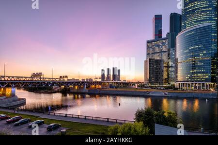 Blick auf den Moskva-Fluss mit Wolkenkratzern und Brücke in der Nacht, Moskau, Russland. Moderne Gebäude im Hintergrund des Sonnenhimmels. Blick auf die Moskau-Ci am Abend Stockfoto