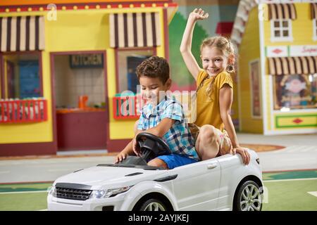 Glückliche Kinder, die ein Elektrospielzeugauto in der Halle fahren. Stockfoto