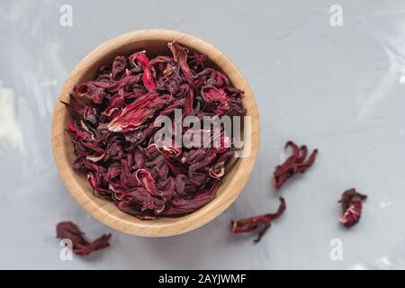 Trockener Tee aus roten Hibiskusblüten, auch Karkade- oder Rosellobst genannt, in Holzschale auf grauem Grund, Konzept des gesunden Getränks. Nahaufnahme Stockfoto