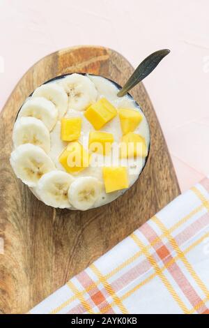 Gelb veganes hausgemachtes Bananen- und Mango-Eis ohne Milchprodukte in Kokosschale mit Löffel, frischen Mango-Stücken und Bananen obenauf, pink Stockfoto