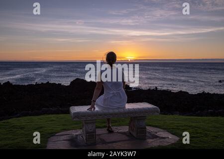 Frau in einem weißen Kleid, die auf einer Steinbank sitzt und den Sonnenuntergang beobachtet Stockfoto