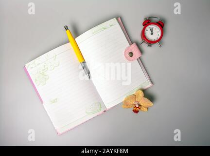 Ein offenes Notizbuch, Stift, Orchideenblume und ein Wecker, der an die Zeit erinnert, stehen auf dem Tisch. Büro. Schulbedarf. Platz für Text. Ansicht von oben Stockfoto