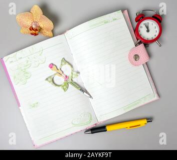 Auf dem Tisch stehen ein offenes Notizbuch, Stift, Orchideenblume, eine Schere und ein Wecker, der an die Zeit erinnert. Büro. Schulbedarf. Platz für Text. Stockfoto