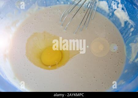 Der Prozess der Zubereitung von Teig für Pfannkuchen. Mit dem Schneebesen in einer blauen Schüssel mit Milch und Mehl auf dem Küchentisch garen. Es stellt sich heraus Stockfoto