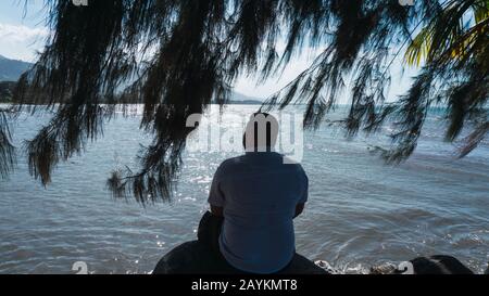 Entlang des Menschen, der auf einem Felsen sitzt, entspannend und mit einem wunderschönen blauen Himmel und Meer reflektiert Stockfoto