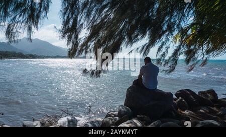 Entlang des Menschen, der auf einem Felsen sitzt, entspannend und mit einem wunderschönen blauen Himmel und Meer reflektiert Stockfoto