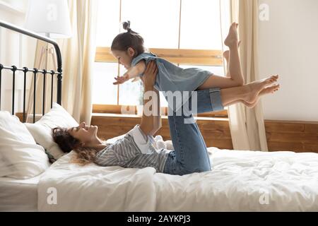 Glückliche junge Mutter hebt kleine Vorschulkindtochter im Schlafzimmer auf. Stockfoto