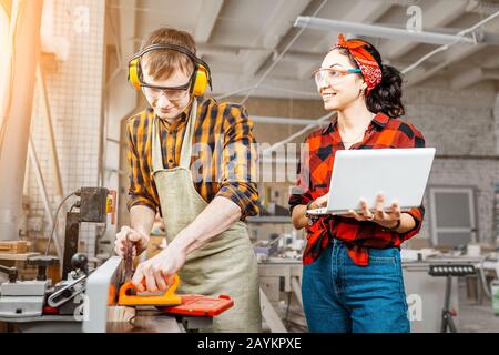 Asiatische Frau mit Laptop und Mann mit Kreissäge, die in einer Fabrik oder Werkstatt arbeiten Stockfoto